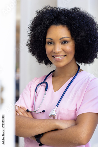 Biracial nurse standing with arms crossed, wearing scrubs at home