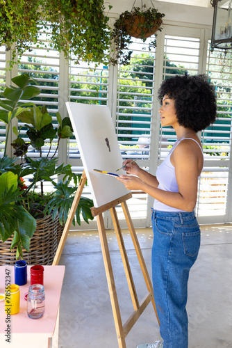 Biracial young woman painting on canvas at home, surrounded by plants
