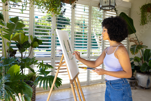Young biracial woman painting on canvas in plant-filled room at home