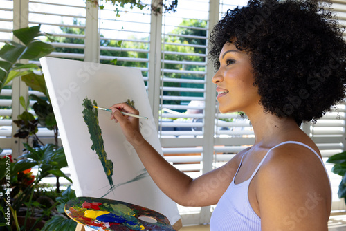 Biracial young woman painting on canvas at home, standing by window
