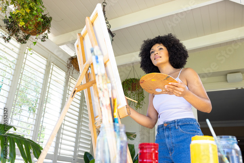 Young biracial woman painting on canvas at home, holding palette