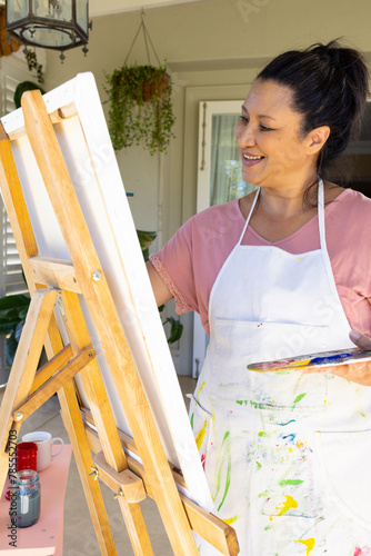 Mature biracial woman painting on easel outdoors at home