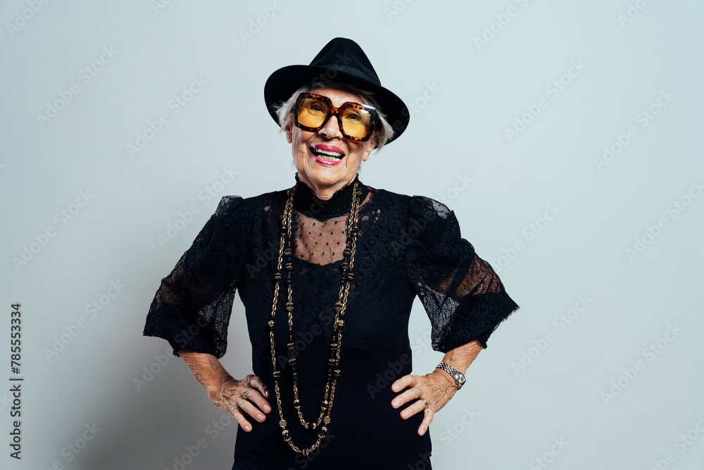 Fototapeta premium image of a beautiful and elegant old influencer woman. Cool grandmother laughing at camera wearing fashionable black outfit with hat and sunglasses 