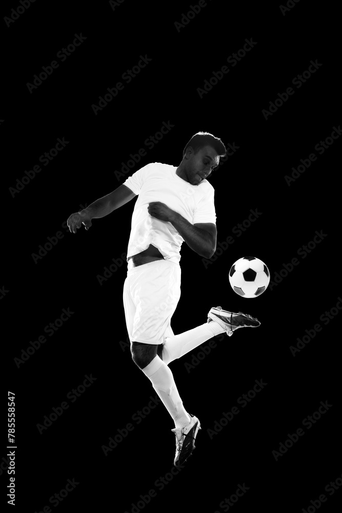 Concentrated young man, soccer player in white uniform training, dribbling ball isolated on black background. Concept of professional sport, game, competition, tournament, action, active lifestyle