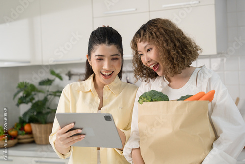 Two women in a kitchen, one holding a tablet and the other holding a grocery bag, both looking surprised and happy.