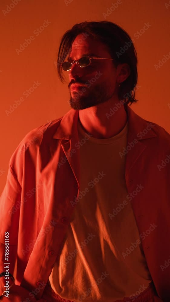 cool young man with retro rectangular sunglasses looking to side, fixing shirt and crossing arms when camera is closing up with red lights on cool young man with retro rectangular sunglasses looking to side, fixing shirt and crossing arms when camera is closing up with red lights on