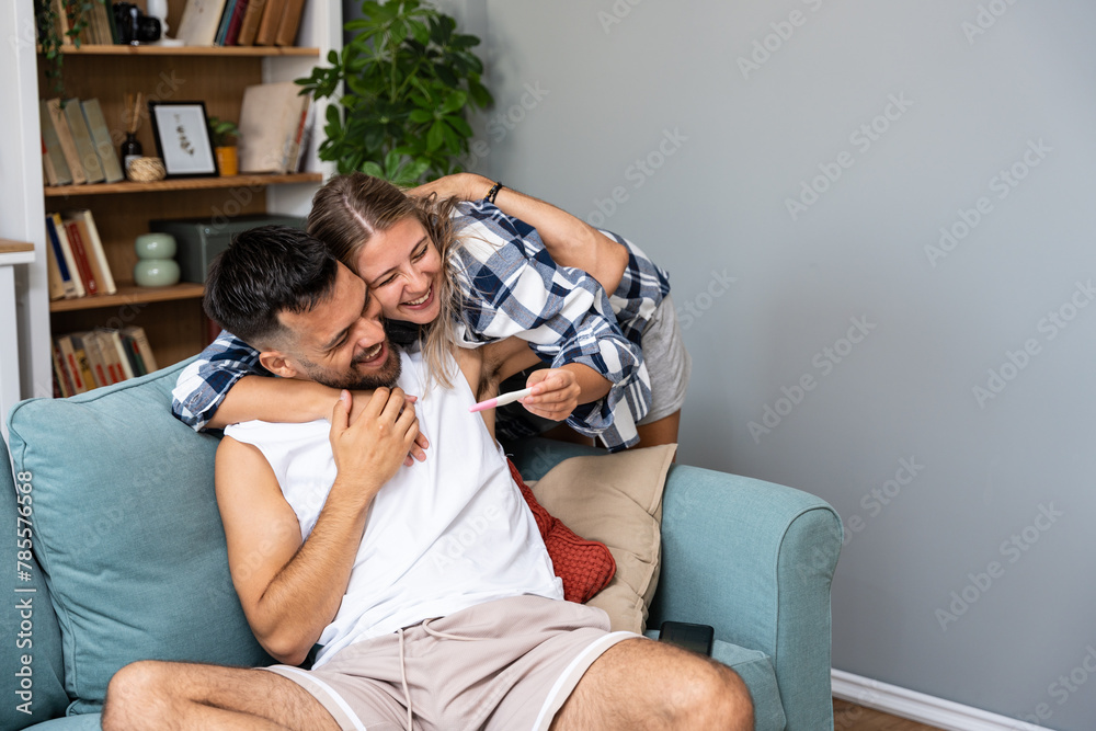 Naklejka premium Young happy couple, pregnant woman showing positive pregnancy test to the man at their apartment. Newlywed couple great news concept.