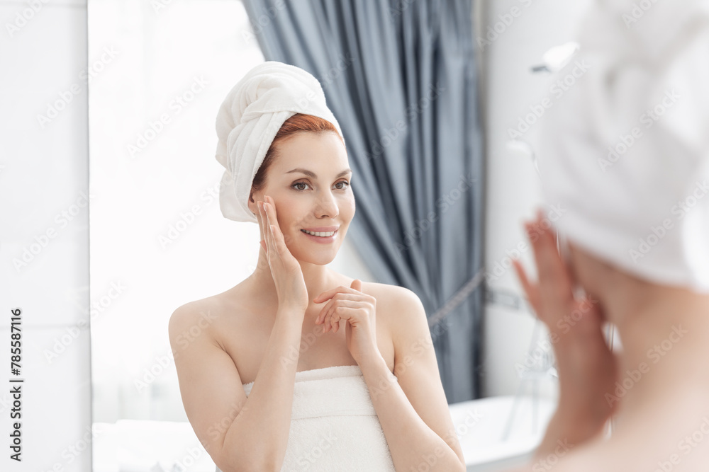 Woman wrapped in towel is examining her face in mirror, focusing on her skin, lips, eyebrows. She smiles at her reflection. Girl with towel on head takes care of her facial skin Woman wrapped in towel is examining her face in mirror, focusing on her skin, lips, eyebrows. She smiles at her reflection. Girl with towel on head takes care of her facial skin