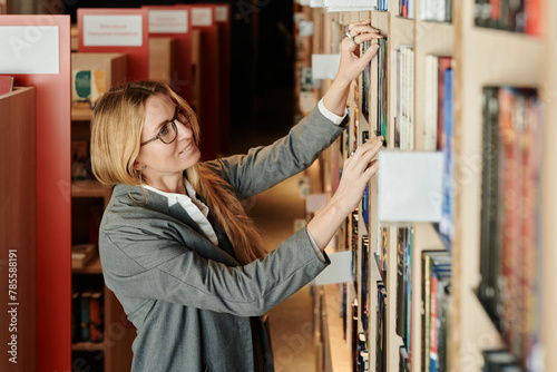 Happy young woman with long blond straight hair standing in front of large stack of shelves in library and looking for book photo