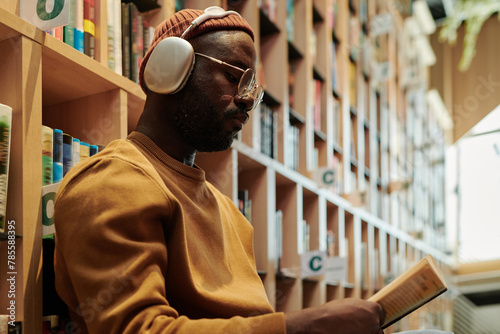 Serious guy in headphones and eyeglasses looking through text in open book while sitting against stacks of bookshelves photo