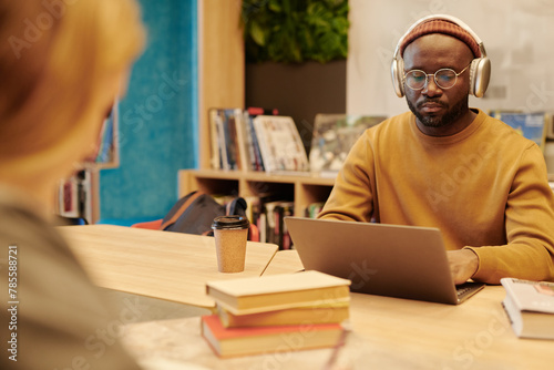 Serious African American guy in eyeglasses, pullover, beanie and headphones networking in front of laptop in library photo