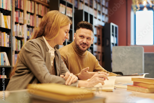 Mature man in quiet luxury attire explaining details of novel plot to blond woman while sitting next to her in reading hall