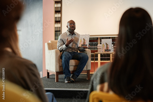 African American author with his new book in hands sitting in armchair in front of public at literature event in modern library photo