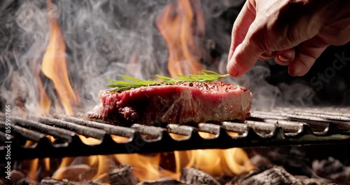 Yellow flames burst out from under grill grate, on which lies juicy piece of steak seasoned with spices, chefs hand gently places sprig of rosemary. Aroma of grilled meat makes your mouth water.