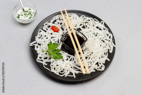 Rice noodles with sauce, bamboo sticks on black plate. Cherry tomatoes in bowl with white sauce on table