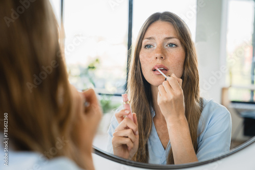 Closeup reflection of young Caucasian woman applying lipstick in front of the mirror at home. Beautiful girl lady female preparing for romantic date, applying cosmetics indoors photo