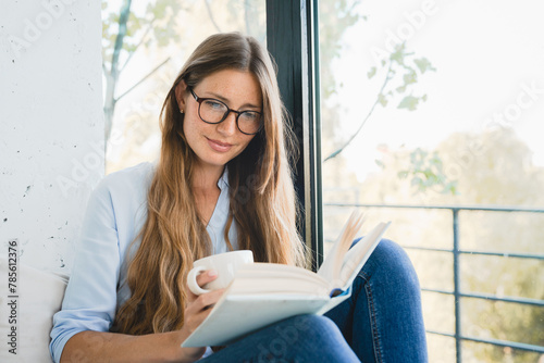 Smart Caucasian woman reading a novel at home by the window. Freelancer student female reading a book, preparing for exam project, doing homework, inspired with the plot photo