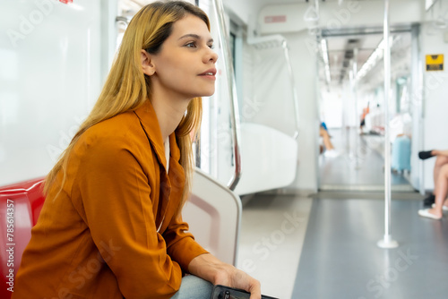 A thoughtful young woman wearing an orange blazer sits inside an empty subway car, looking into the distance, surrounded by urban tranquility.