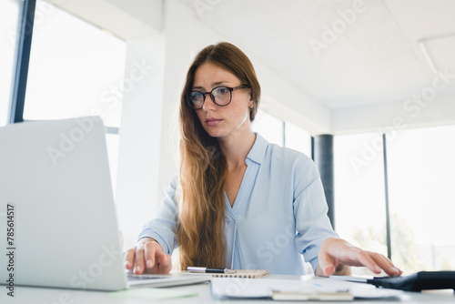 Young Caucasian woman counting percent of budget using laptop and calculator at office desk. Female freelancer typing, preparing report, paying loan debt bills overdue