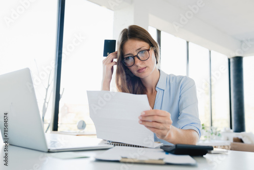 Confused young freelancer reading official letter with loan debt mortgage papers at office desk, Sad depressed focused girl having problems issues at workplace with billing overdue photo