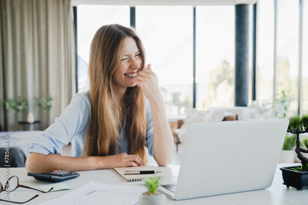 Smiley young woman calculating funds and laughing while using laptop at home office. Caucasian freelancer in positive mood paying domestic bills, loans, mortgage, insurance Smiley young woman calculating funds and laughing while using laptop at home office. Caucasian freelancer in positive mood paying domestic bills, loans, mortgage, insurance