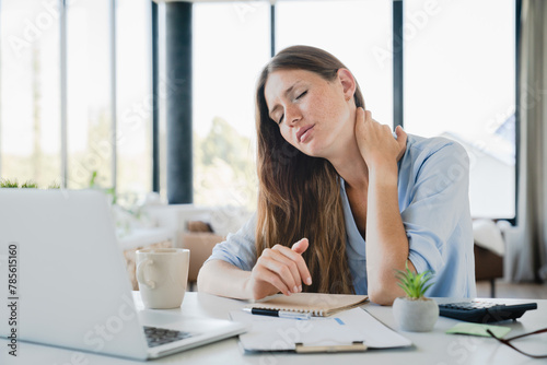 Tired young woman suffering from neck pain while working on laptop at home desk. Freelancer manager office employee rubbing muscles for ache pain relief photo