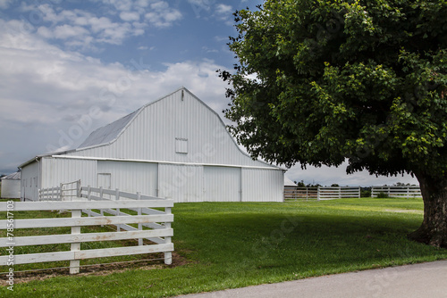 White Barn with a White Fence.  A large white barn stands prominent against a cloudy sky, complemented by a lush green tree to its right. 