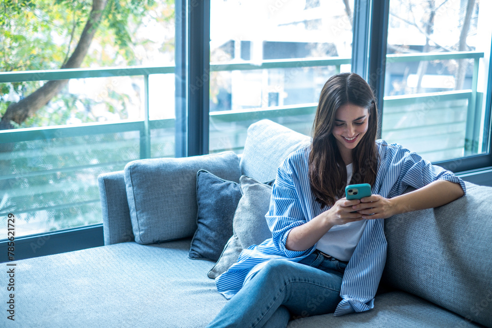 Happy girl scrolling and checking social media while holding smartphone at home. Smiling young brunette woman using mobile phone app to play games, shop online, order delivery while relaxing on sofa. Happy girl scrolling and checking social media while holding smartphone at home. Smiling young brunette woman using mobile phone app to play games, shop online, order delivery while relaxing on sofa.