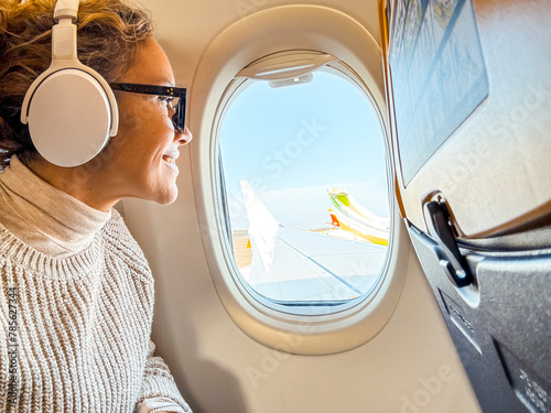 Canvas Print Smiling passenger with Bluetooth noise-canceling headphones looking out of the airplane's porthole using the on-board Wi-Fi Internet connection