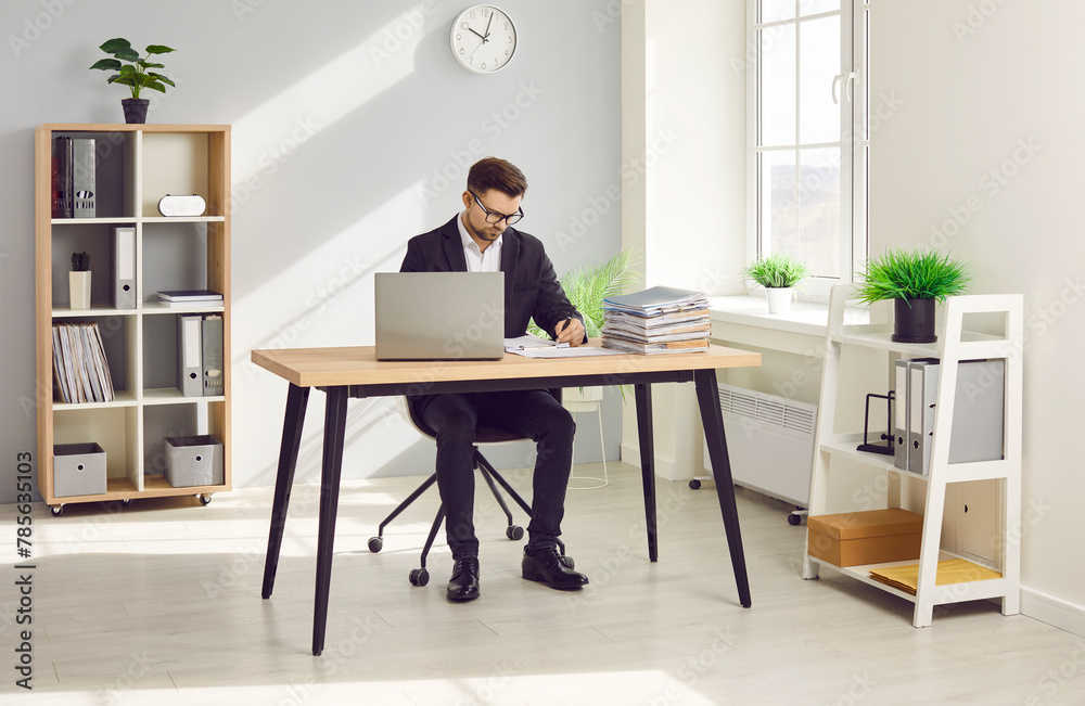 Businessman working in office with laptop and papers on desk. Focused serious man in glasses and formal wear sitting at desk analyzing data and writing notes at workplace Businessman working in office with laptop and papers on desk. Focused serious man in glasses and formal wear sitting at desk analyzing data and writing notes at workplace