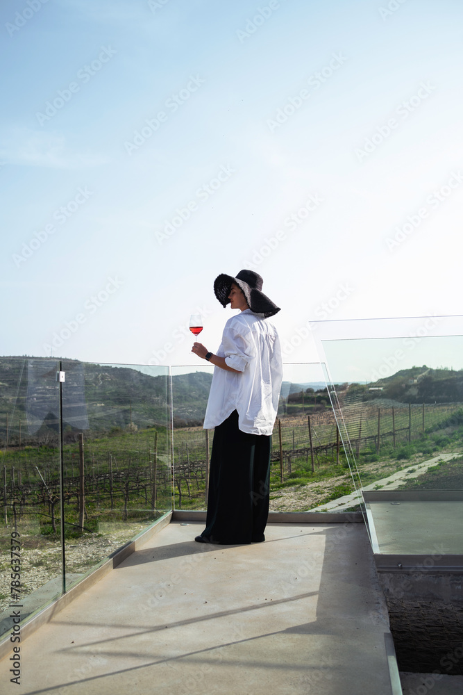 Fototapeta premium Woman in elegant attire and a wide-brimmed hat enjoys a glass of rose wine while overlooking a sprawling vineyard