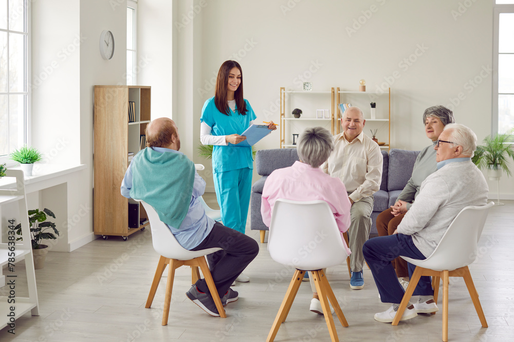 Happy senior people at retirement home sitting in circle in living room and having discussion with young girl nurse, carer, therapist, psychologist or counselor with clipboard. Group therapy concept