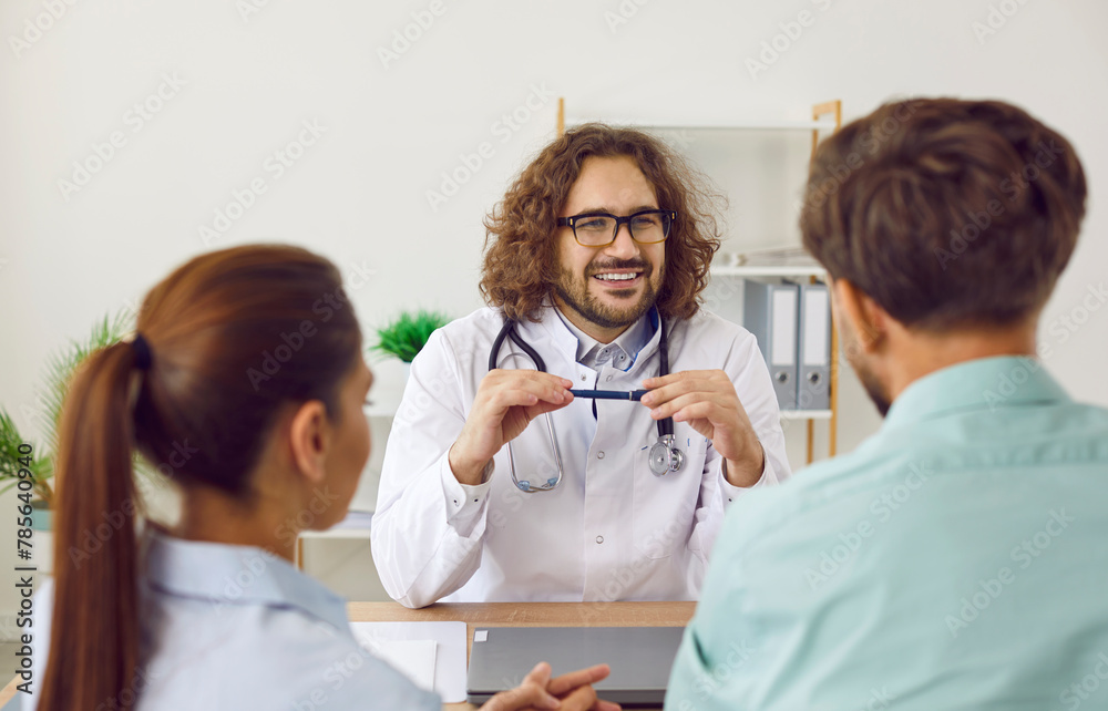 Obraz premium Cheerful friendly male doctor talking to patients about IVF treatment in his clinic. Happy smiling man in white medical coat and glasses sitting at table and looking at young family couple