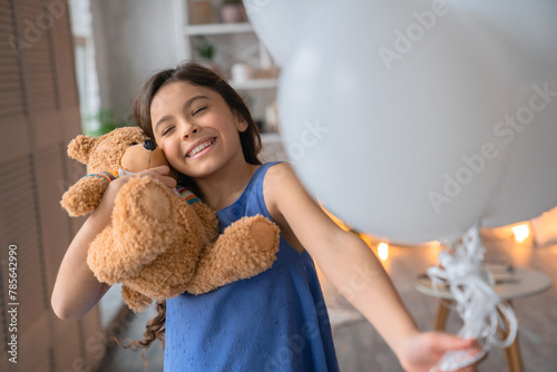 Close up portrait of smiling cheerful pretty girl with long wavy brown hair hugging her teddy bear toy with white colorful air balloons in her hand, closing eyes and toothy smile. Blure background