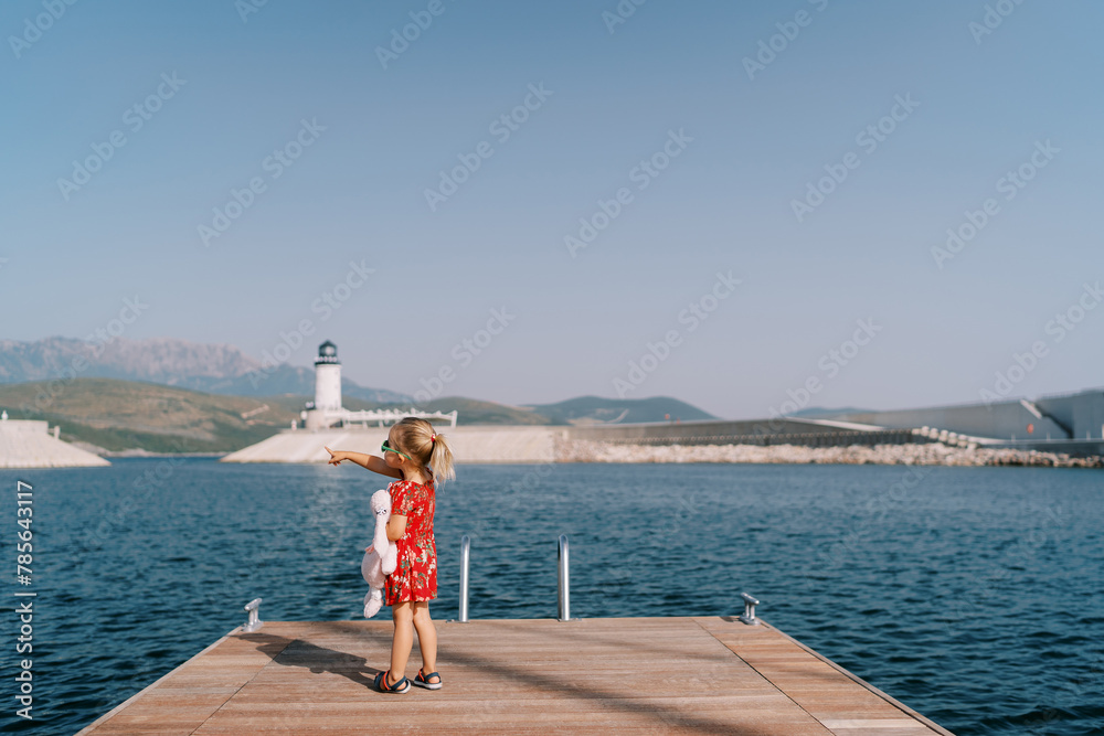 Little girl with a toy rabbit stands on the pier and points to the mountains. Back view