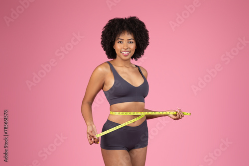 Smiling African American lady measuring waist on pink background