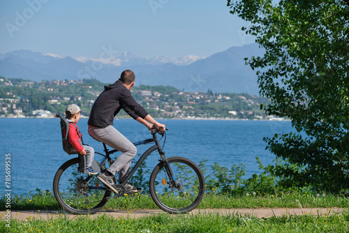 a man with a child on a mountain bike in motion with a lake in the background, mountain peaks in the snow.