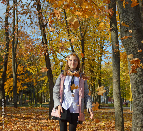 beautiful, cheerful girl on a walk in the autumn park. a child walks through fallen maple leaves and enjoys the good weather and warm autumn sun. from above, yellow maple leaves fall on the head