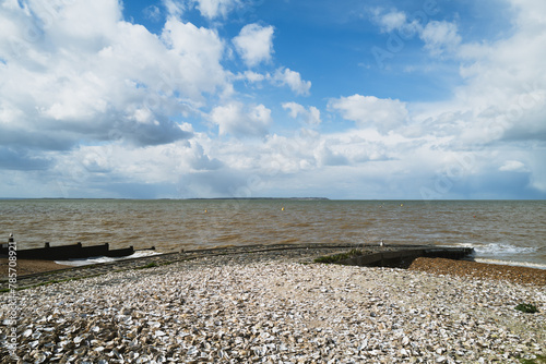 Oyster shells on Whitstable beach next to a slipway with a wooden groyne to the left. The clouds are dramatic. The isle of Sheppey can be seen on the horizon.