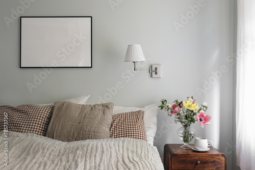 Elegant bedroom. Tulips, cherry tree blossoms bouquet in glass vase. Wooden night stand. Cup of coffee. Blank black picture frame mockup. Lamp, pleated shade on mint wall. Checkered pillows.