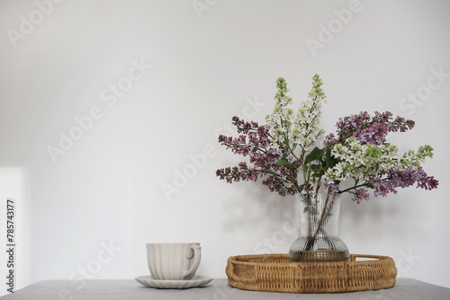 Moment of tranquility with cup of coffee, wicker tray. Floral bouquet of blooming purple and white lilacs branches,glass vase. Springtime breakfast scene, table. White wall background, selective focus © tabitazn