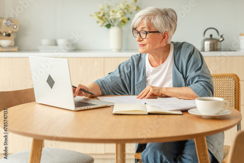 Online education courses webinar concept. Middle aged senior woman using laptop computer writing notes. Focused mature old woman enjoying studying online from home written records doing online work