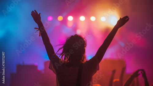 A woman enjoying a concert, lifting her arms in the air with the stage lights casting vibrant hues over her. Shallow depth of field, blurred background