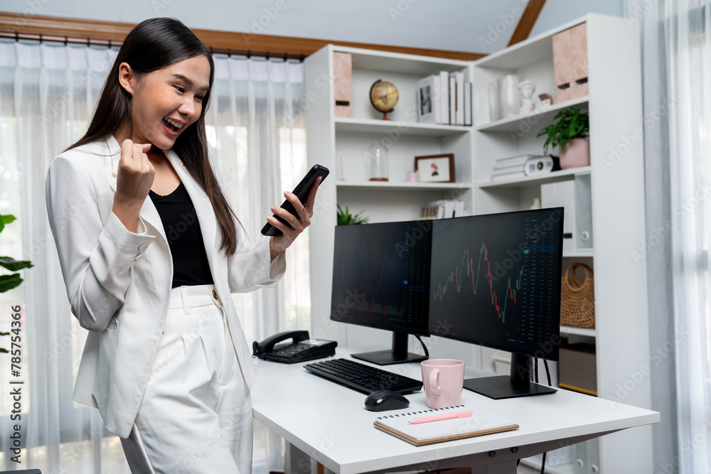 Successful young Asian businesswoman earning high profit raising fist up, holding smartphone in exchange stock market's financial technology with dynamic business graph at modern office. Stratagem.