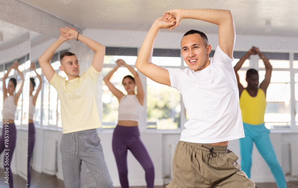 Young Asian male instructor in casual sportswear enthusiastically guiding diverse class through zumba routine in fitness studio, emphasizing fun and rhythm..