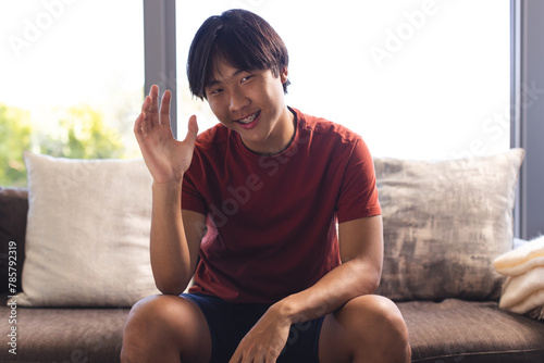 Asian teenage boy sitting, having a video call at home, waving at the camera, wearing a red shirt