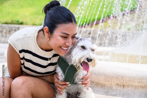 Cheerful young woman in a casual outfit cuddling with her adorable schnauzer beside a water fountain