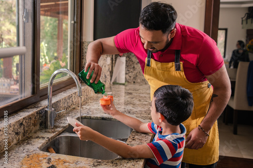 Obraz na plátně Latin father helps his son put soap on the sponge to wash the dishes
