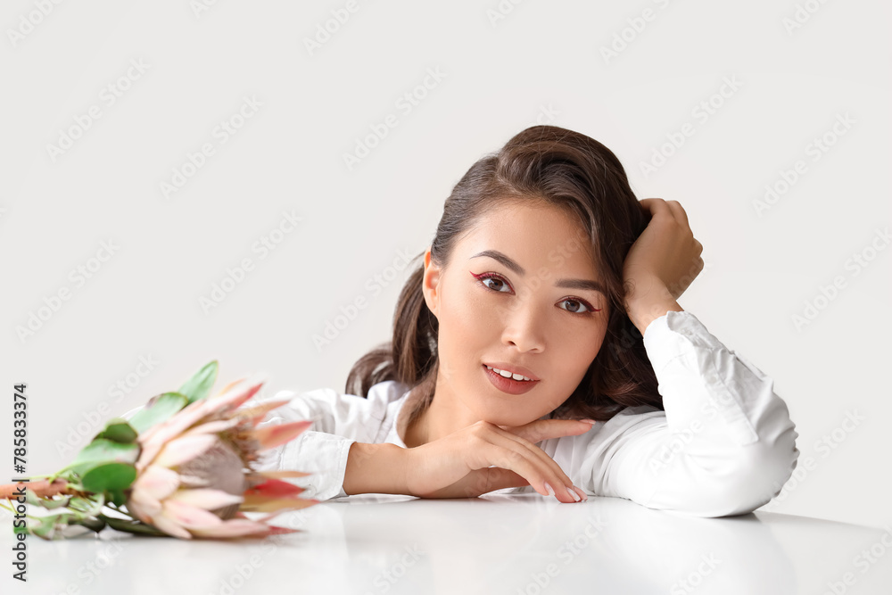 Young Asian woman with beautiful protea flower at table near white wall