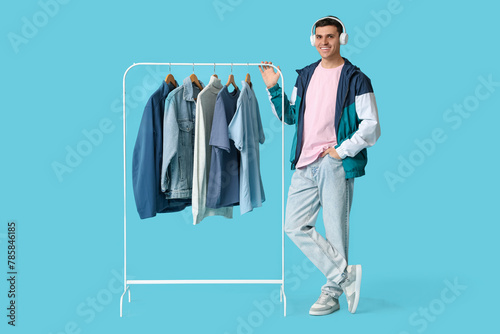 Handsome young man in headphones and rack with stylish clothes on blue background